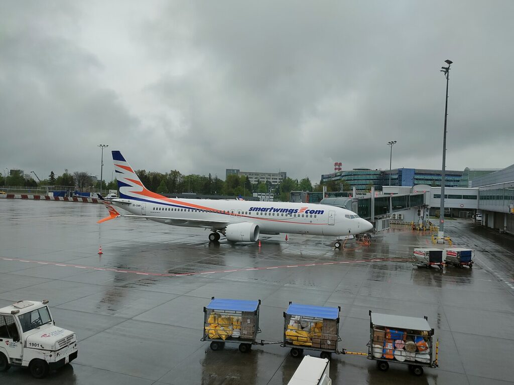 Boeing 737 MAX 8 společnosti Smartwings v modro-oranžových barvách na stojánce pražského letiště za deštivého počasí, vedle zavazadlové vozíky